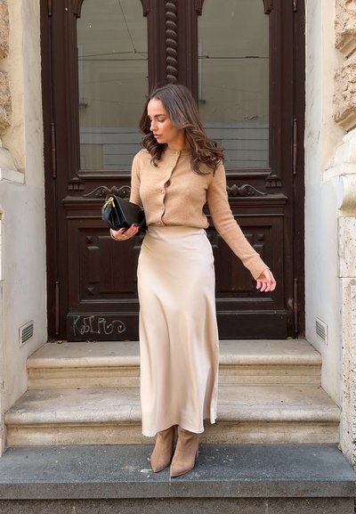 Cardigan de lana beige con botones, falda larga de satén color crema y botas de tobillo color marrón. Sosteniendo un pequeño bolso negro, de pie en escalones de piedra.