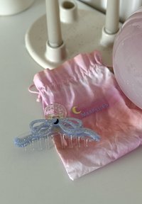 Clip per capelli in acrilico di colore azzurro chiaro con strass, design a fiocco, denti trasparenti, posizionato su un sacchetto con cordoncino in gradiente rosa e bianco.