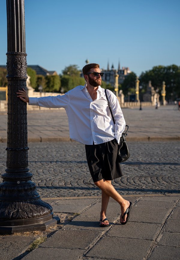 Hombre con camisa blanca, pantalones cortos negros, gafas de sol y chanclas apoyado en un farol ornamentado en una plaza abierta y soleada con suelo empedrado.