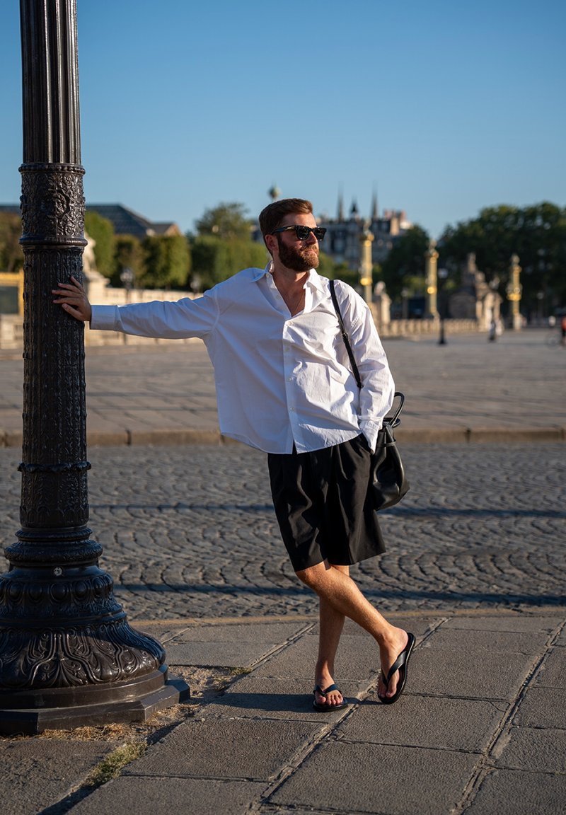Homme en chemise blanche, short noir, lunettes de soleil et tongs appuyé contre un lampadaire orné dans une place ouverte et ensoleillée avec un sol pavé.