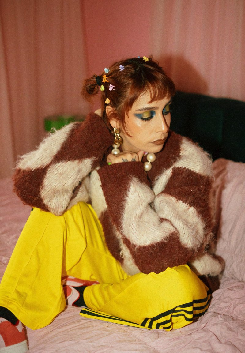 Personne avec des barrettes colorées, un maquillage des yeux audacieux, un pull oversize et un pantalon jaune assise en tailleur sur un lit avec un fond rose.