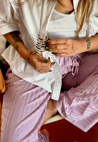 Weißes Hemd mit Kragen, hellrosa gestreifte Hose, lange grüne Nägel, hält eine Haarklammer, sichtbare Tattoos an den Armen, Holzstuhl.