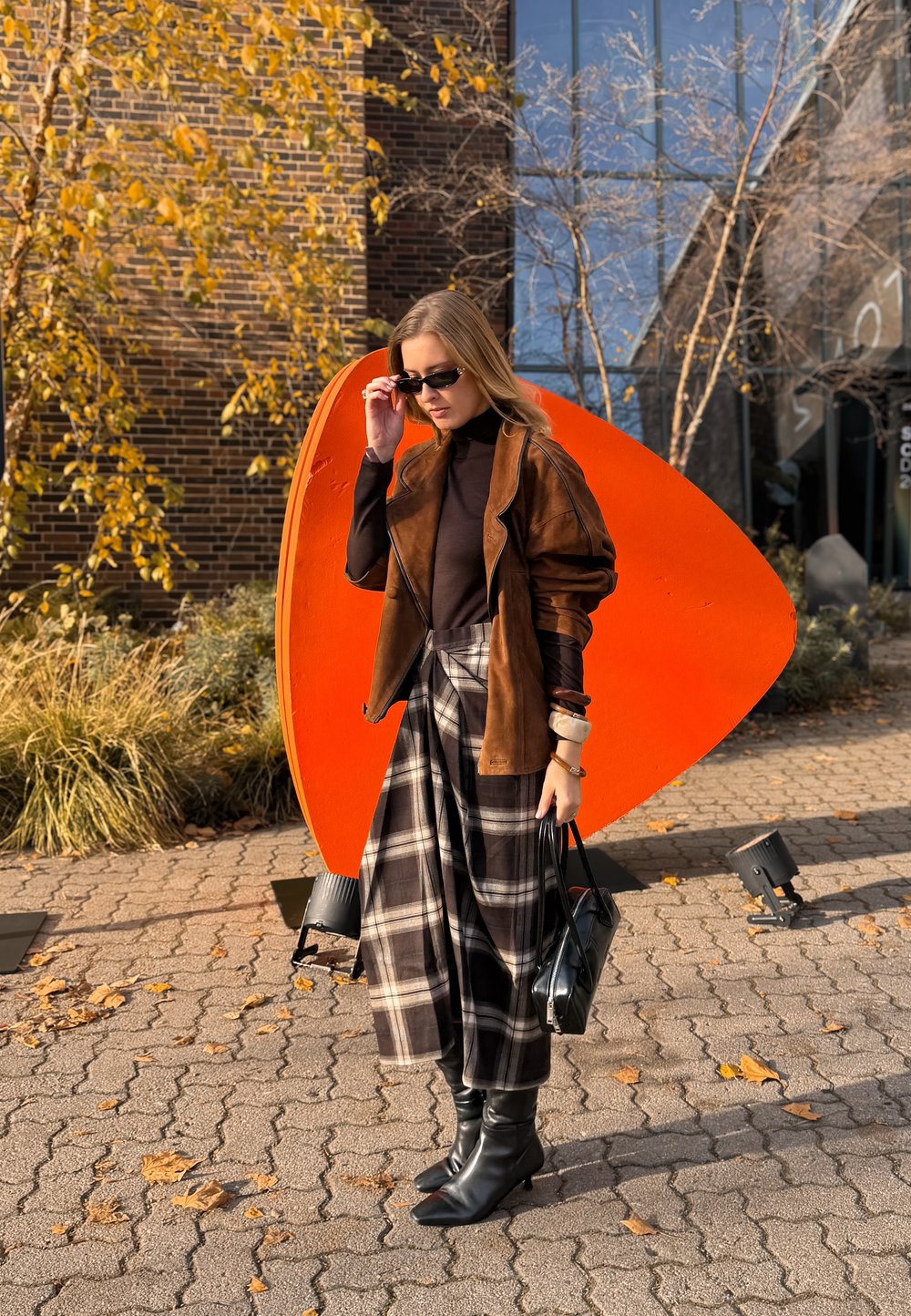 Hnedá semišová bunda cez čierny rolák, v kombinácii s károvanou maxi sukňou. Čierne čižmy po kolená, v ruke čierna kabelka. V pozadí oranžová socha.