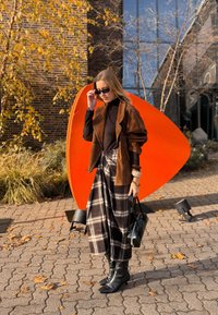 Hnedá semišová bunda cez čierny rolák, v kombinácii s károvanou maxi sukňou. Čierne čižmy po kolená, v ruke čierna kabelka. V pozadí oranžová socha.