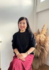 Sweat-shirt noir, jupe bordeaux, et bracelet doré. Cheveux longs et fonc és, assise contre un mur clair avec un vase de feuillage sec.