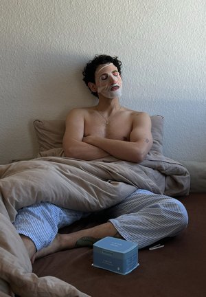 Maschera facciale applicata, persona seduta con capelli ricci, che indossa pantaloni del pigiama a righe. Biancheria da letto beige e un contenitore di prodotti per la cura della pelle di colore blu chiaro nelle vicinanze.