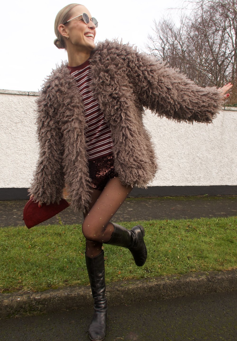 Manteau marron pelucheux surdimensionné, chemise à rayures, short à paillettes, collants transparents avec des accents dorés, et bottes noires montantes jusqu'aux genoux. Pochette rouge à la main.