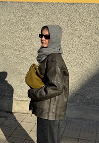Cachecol cinza e jaqueta de couro escura, segurando uma bolsa amarela texturizada. Apresenta formas angulares e um brilho sutil. Fundo urbano neutro.