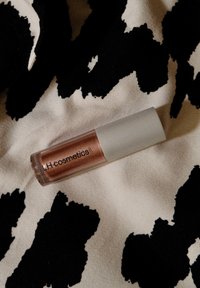Lipgloss i klar beholder med en sølvfarvet hætte, der har en metalisk bronze nuance. Baggrunden har et sort-hvidt cowprint mønster.