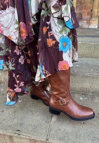 Bottes hautes en cuir marron jusqu'aux genoux avec un talon épais et un détail de boucle. Associées à une robe à volants à fleurs avec des motifs de fleurs bleu vif et roses.