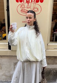 Jachetă albă plisată cu guler înalt și fermoar. Fustă albă asortată, ținând o cană de cafea. Geantă maro cu model. Fundalul prezintă un logo roșu de cafea.