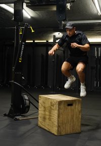 Άνδρας με μαύρη αθλητική ενδυμασία και καπέλο εκτελεί άλμα πάνω από ξύλινο πλαίσιο πλαστικής άσκησης σε γυμναστήριο με εξοπλισμό γυμναστικής.