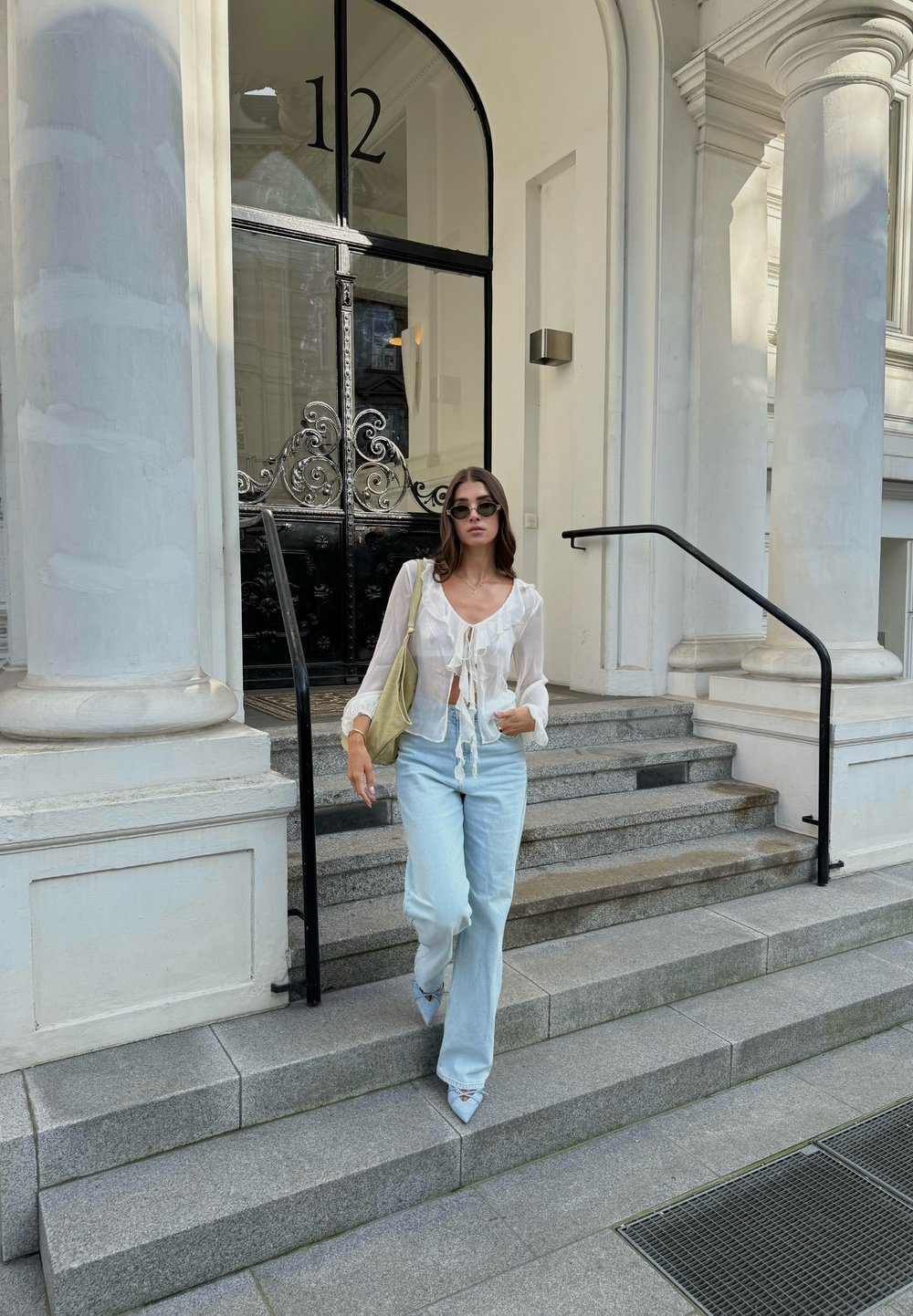 Blusa leve, branca suja, com detalhe em laço, combinada com calças jeans de perna larga em azul claro e sapatos de bico fino de cor clara. Acento de bolsa verde.