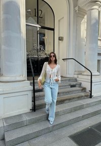 Blusa leve, branca suja, com detalhe em laço, combinada com calças jeans de perna larga em azul claro e sapatos de bico fino de cor clara. Acento de bolsa verde.