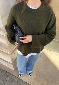 Maglione oversize verde oliva con finitura testurizzata; abbinato a jeans a gamba larga blu chiaro e una pochette intrecciata nera con design annodato.