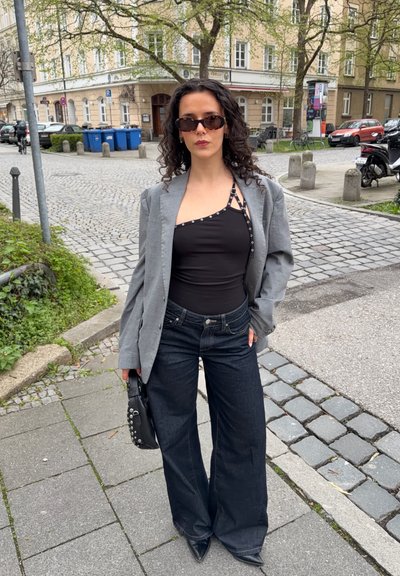Mujer con cabello rizado que lleva gafas de sol, top negro asimétrico, blazer gris, vaqueros de pierna ancha y sostiene un bolso negro con tachuelas en la acera de la ciudad.