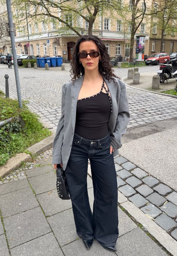 Γυναίκα με σγουρά μαλλιά που φοράει γυαλιά ηλίου, ασύμμετρο μαύρο τοπ, γκρι σακάκι, τζιν παντελόνι με φαρδιά πόδια και κρατάει μαύρη τσάντα με τρουκς σε πεζοδρόμιο της πόλης.