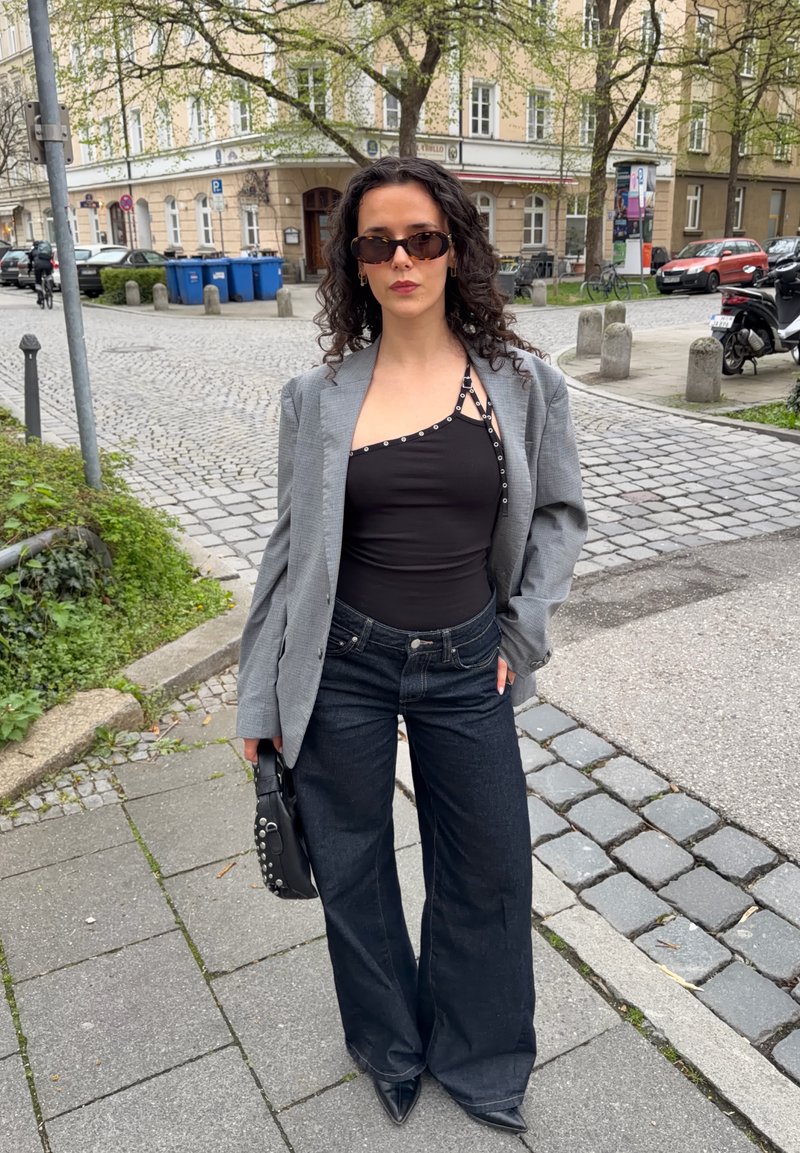 Γυναίκα με σγουρά μαλλιά που φοράει γυαλιά ηλίου, ασύμμετρο μαύρο τοπ, γκρι σακάκι, τζιν παντελόνι με φαρδιά πόδια και κρατάει μαύρη τσάντα με τρουκς σε πεζοδρόμιο της πόλης.