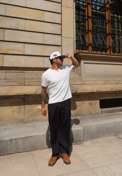 Hombre con camisa blanca de manga corta, pantalones negros a rayas, botas marrones y gorra blanca que le tapa los ojos, de pie junto a un edificio de piedra con ventanas con rejas.