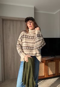 Femme portant un pull beige à motifs et une casquette, tenant une écharpe verte, debout à l'intérieur à côté d'un meuble en bois avec une télévision.