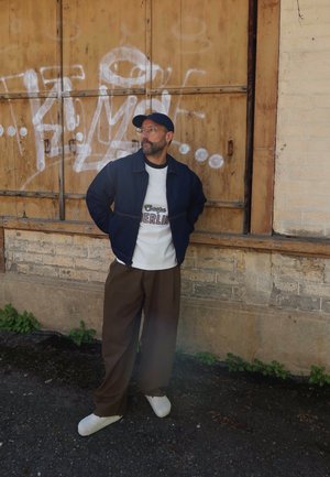 Hombre con gafas, gorra, chaqueta azul marino, camiseta blanca con la palabra "Berlin" y pantalones marrones, de pie junto a una puerta de madera cubierta de grafitis y una pared de ladrillos.