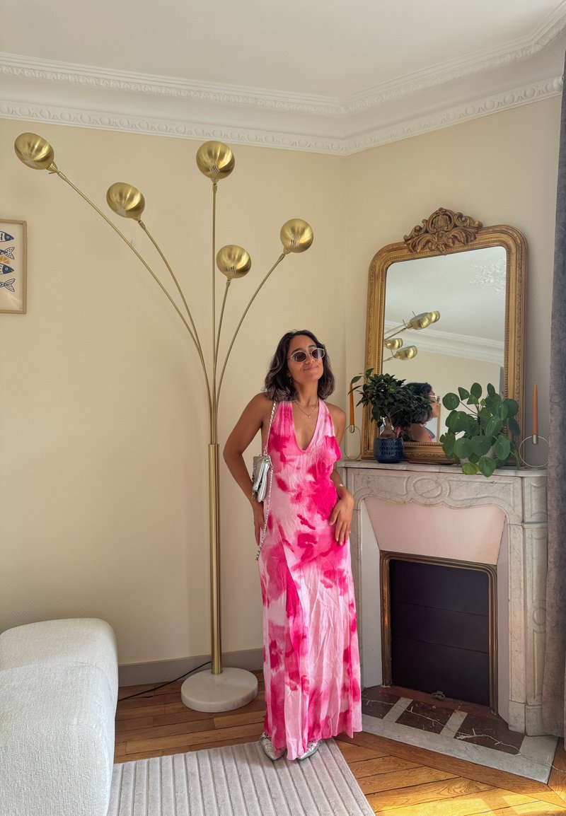 Femme portant une robe tie-dye rose et blanche, des lunettes de soleil et un sac argenté, se tient à côté d'une lampe sur pied dorée et d'une cheminée en marbre avec des plantes et un miroir.