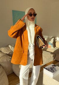 Brun oversized blazer i semsket skinn med to fremre lommer, brukt over en hvit bluse. Tilbehøret inkluderer solbriller og en teksturert clutch.