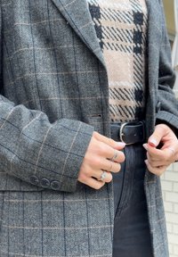 Hall hallhoundsuuriga bleiser struktureeritud lõikega, millel on üks nööp ja detailirohked varrukaservad, kantud beeži ja musta ruudulise kampsuni peal.