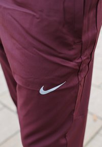 Pantalon de sport marron en tissu lisse, avec un logo Nike argenté sur le côté gauche et une coupe épurée avec des coutures latérales.