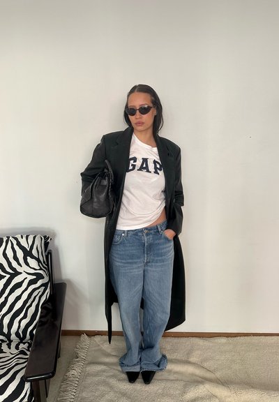 Mujer con abrigo de cuero negro, camiseta blanca de GAP, jeans azules, gafas de sol negras y sosteniendo un bolso negro, de pie en el interior cerca de una silla con patrón de cebra.