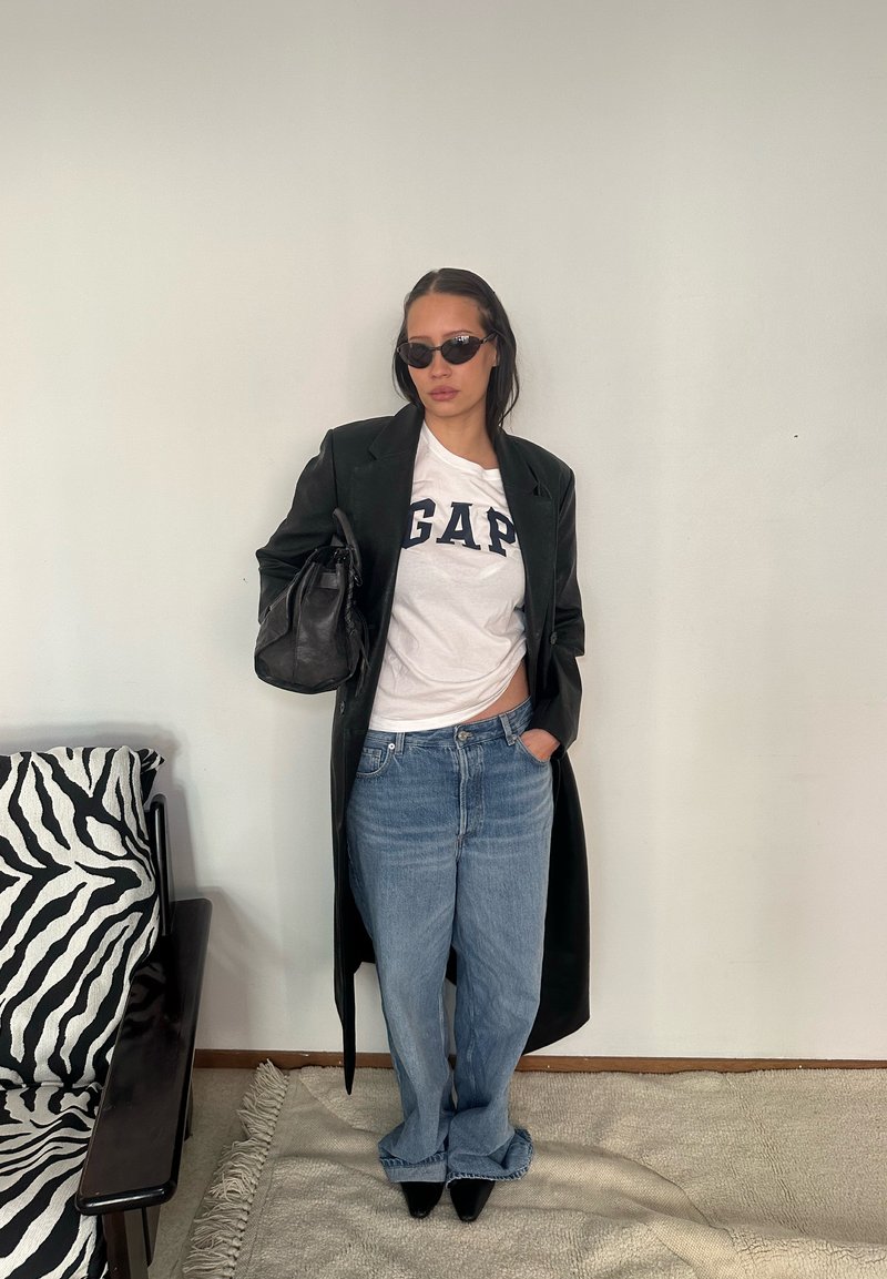 Femme portant un manteau en cuir noir, un t-shirt blanc GAP, un jean bleu, des lunettes de soleil noires, tenant un sac à main noir, debout à l'intérieur près d'une chaise à motif zèbre.