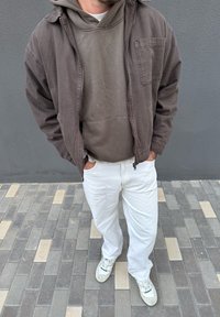 Man wearing a dark brown jacket over a brown hoodie, white trousers, and white trainers standing on a grey and beige tiled pavement.