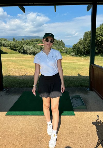 Camisa polo de cuello blanco, falda negra y zapatillas blancas. Los accesorios incluyen gafas de sol y una gorra verde. Césped y montañas de fondo.