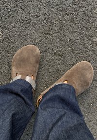 Bruna mockasklackar med silverfärgad metallsmyckning och sidoutskärningar. Bärs med mörkblå jeans byxor på en texturerad asfaltyta.