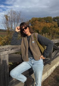 Colete de polar castanho sobre um suéter escuro, jeans de lavagem clara e botas castanhas. Acessórios incluem óculos de sol pretos e uma bolsa verde-oliva.