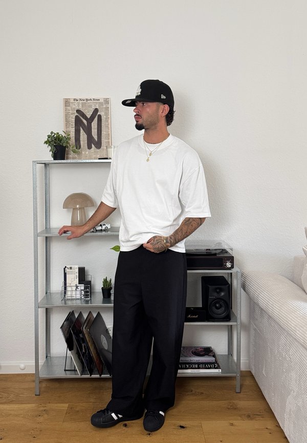T-shirt blanche surdimensionnée, pantalon large noir, baskets noires. Scène intérieure avec étagère en métal, lampe, disques vinyles et plantes en petits pots.