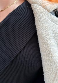 Gros plan d'une personne portant un pull noir côtelé, une veste polaire blanche, un collier en or et des mèches de cheveux rouges visibles.