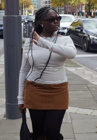 Camisola de manga comprida às riscas, mini saia de camurça castanha, leggings pretas, óculos de sol e cabelo trançado; segurando um guarda-chuva com uma mão.