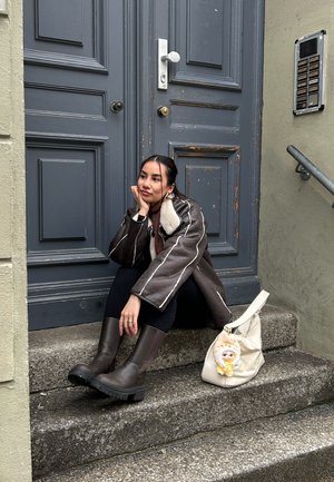 Joven con chaqueta de borrego marrón y botas, sentada en unos escalones de piedra junto a una puerta azul desgastada, con una bolsa blanca a su lado.