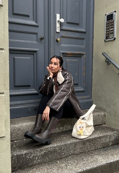 Joven con chaqueta de borrego marrón y botas, sentada en unos escalones de piedra junto a una puerta azul desgastada, con una bolsa blanca a su lado.