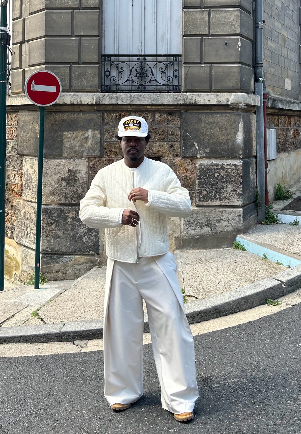 Jachetă bomber cu matlasare albă, pantaloni crem cu croială lejeră și o șapcă cu model. Stând la colțul unei străzi cu piatră texturată și paviment.