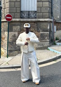 Jachetă bomber cu matlasare albă, pantaloni crem cu croială lejeră și o șapcă cu model. Stând la colțul unei străzi cu piatră texturată și paviment.