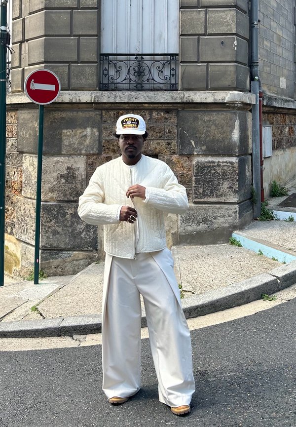 Chaqueta bomber blanca acolchada, pantalones crema de corte relajado y una gorra con estampado. De pie en una esquina de la calle con piedra texturizada y pavimento.
