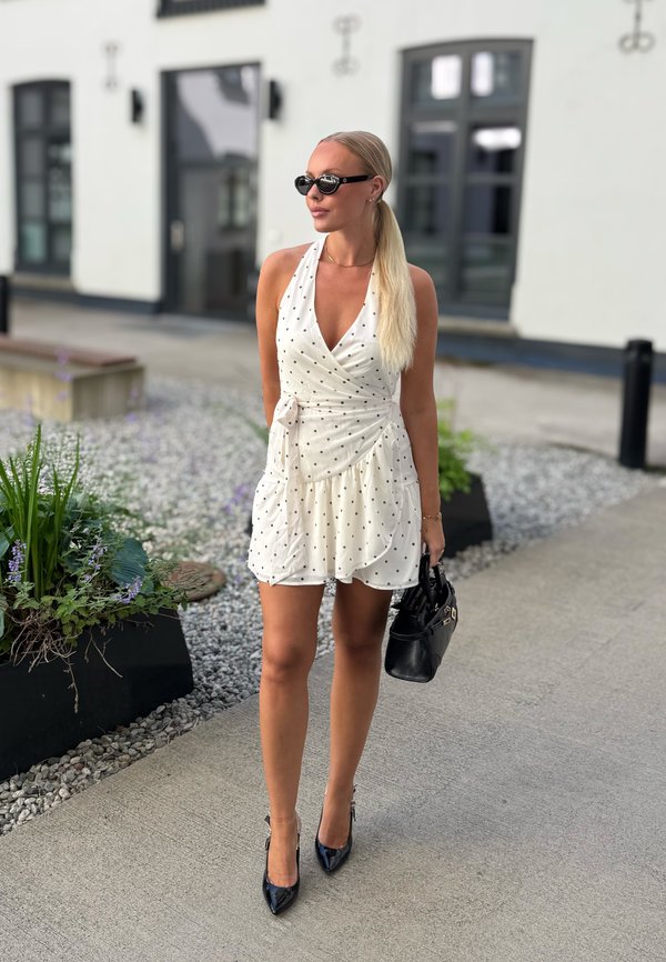 Žena s dlhými blond vlasmi v bielych šatách s bodkami a čiernych lodičkách kráča vonku, nesie čiernu kabelku a nosí tmavé slnečné okuliare.