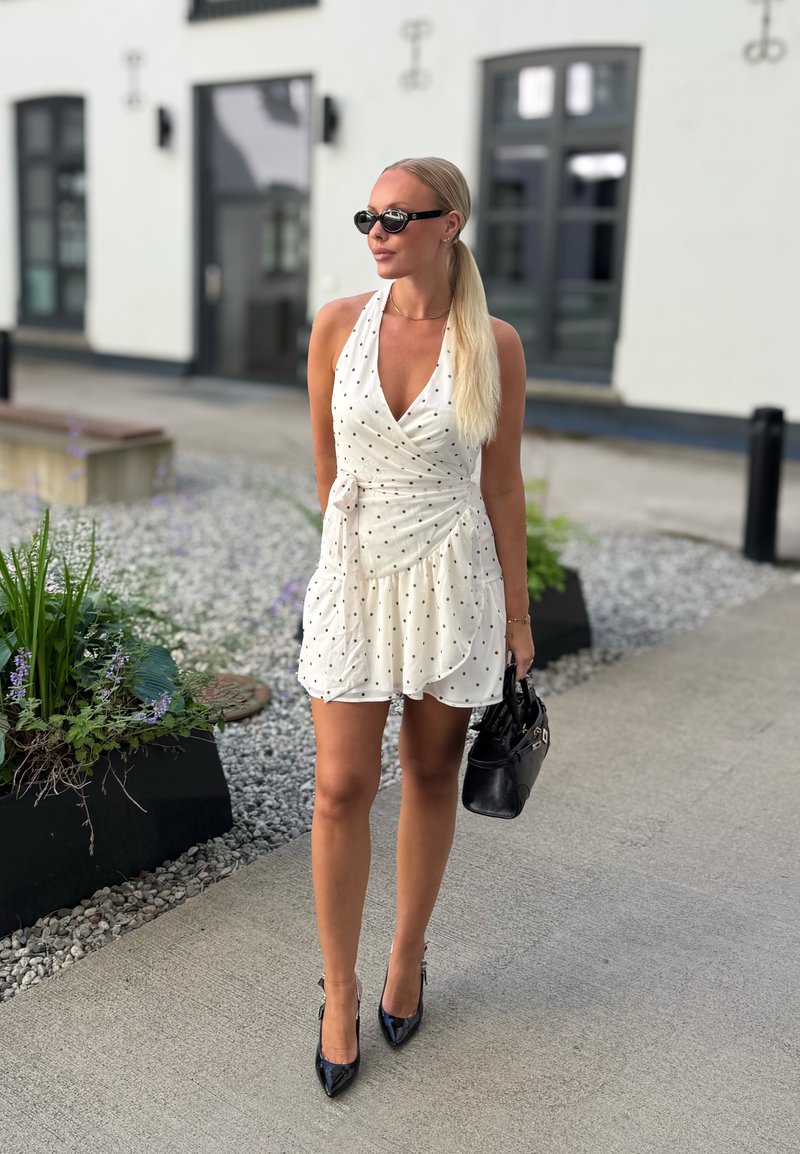 Femme aux longs cheveux blonds dans une robe blanche à pois et des talons noirs marchant en plein air, portant un sac à main noir et des lunettes de soleil foncées.