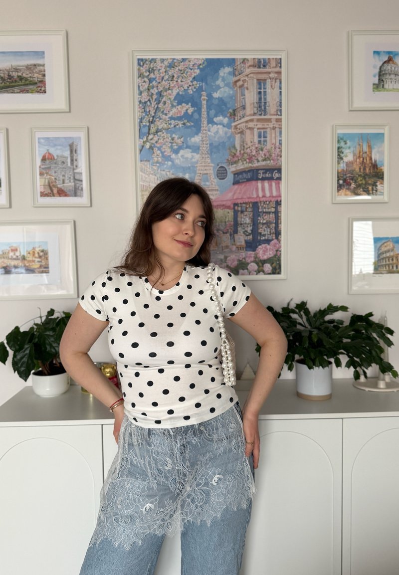 Femme en chemise blanche à pois et jean en dentelle, debout les mains sur les hanches devant un mur orné de peintures encadrées représentant des paysages urbains et de plantes.