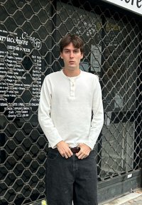 Chemise blanche à manches longues de style henley avec un col boutonné, associée à un pantalon en denim foncé. En arrière-plan, une grille métallique et du texte sur une vitrine de magasin.