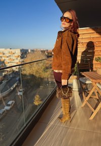 Giacca oversize in suede marrone, leggings bordeaux, stivali color tan al ginocchio e una borsa soffice con catena. Ambiente esterno luminoso con cielo sereno.