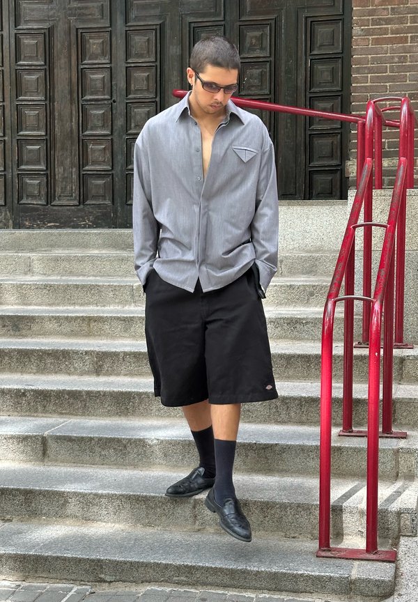 Homme aux cheveux courts portant des lunettes de soleil, une chemise grise ample, un short noir et des chaussures habillées descend des marches en pierre avec des rampes rouges.