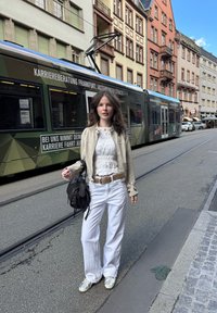 Fehér, bő szárú nadrág, fehér textúrált felső, világos bézs dzseki, fekete táska és ezüst sportcipő. Városi környezet, háttérben villamossal.